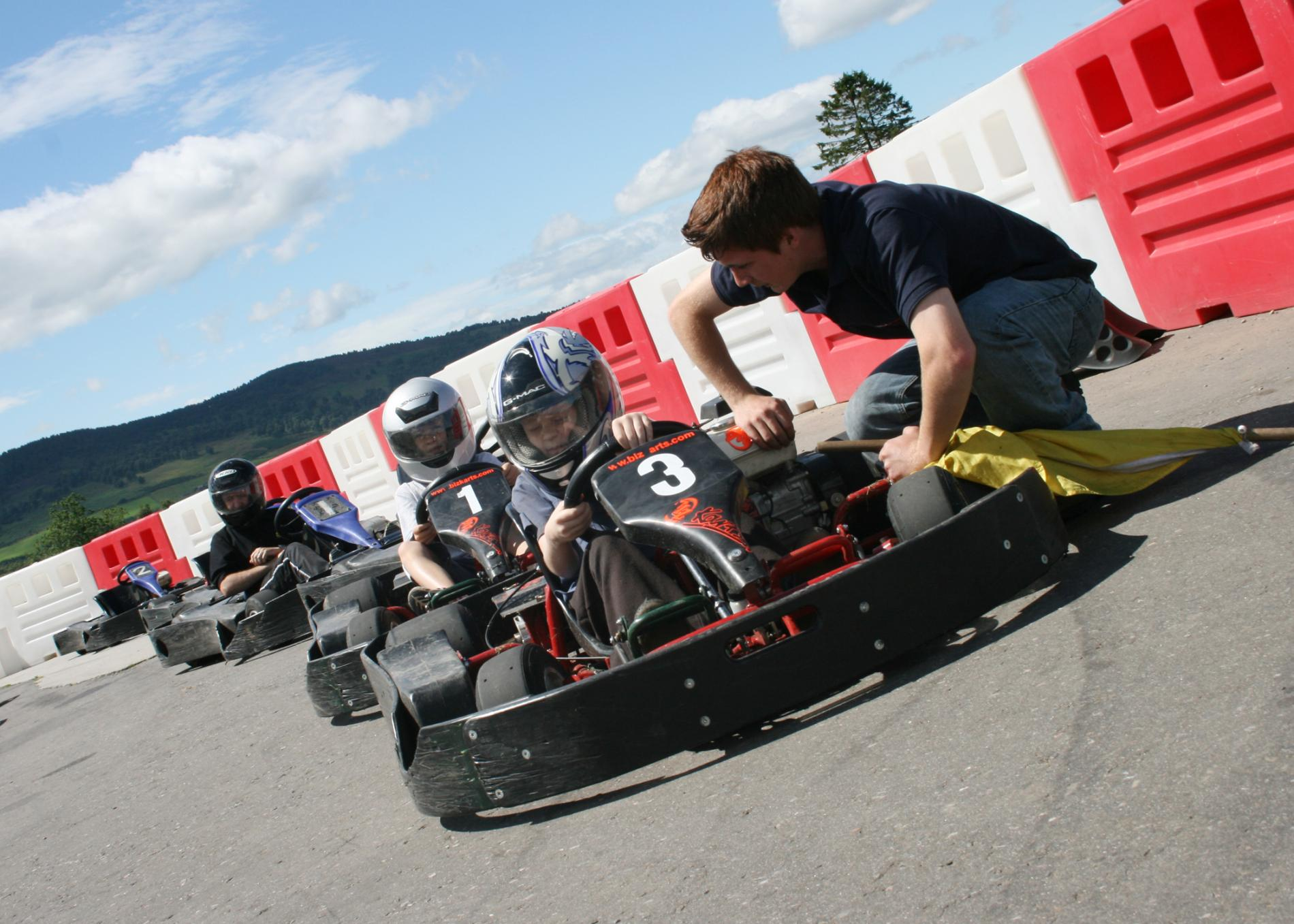 A group of four go-kart racers on a sunny day are lined up beside red and white barriers. A man kneels, assisting the front racer with helmet number 3.